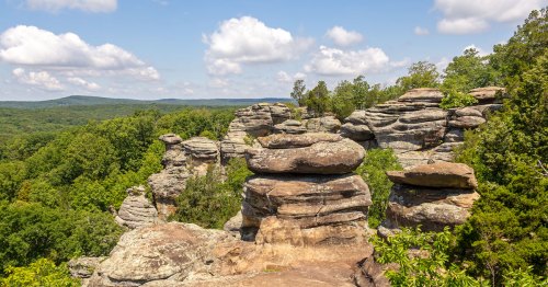 Rock formations tower over a green forest with a blue sky.