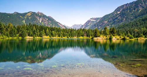Serene alpine lake with crystal-clear water reflecting evergreen forests and rugged mountain peaks beneath a bright blue summer sky.