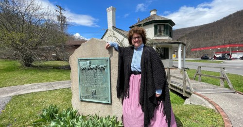 a woman dressed in a long pink skirt, blue long-sleeved shirt, and a black shawl stands outside the lavale toll gate house in maryland