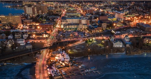 It's nighttime as we're positioned over a brightly lit town with a wide and mostly empty bridge moving over a river.