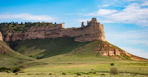 A towering sandstone bluff rises dramatically above a green grassy slope under a blue summer sky at Scotts Bluff National Monument in Nebraska.