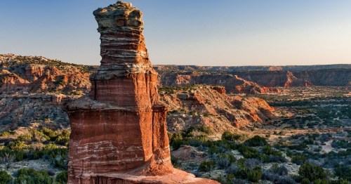 Towering red rock formation known as The Lighthouse rises above rugged canyon walls and desert vegetation in Palo Duro Canyon State Park, showcasing one of Texas’ most iconic scenic viewpoints under a clear blue sky.