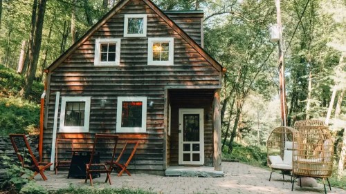 A rustic wooden cabin surrounded by trees, featuring a patio with chairs and a cozy atmosphere.