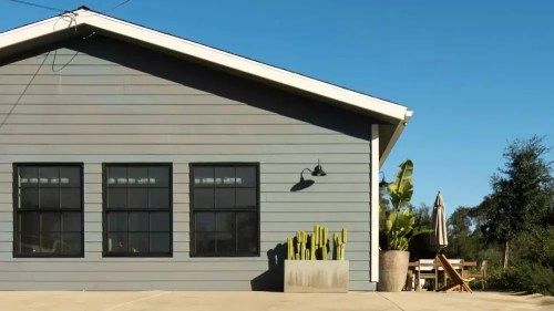 A modern gray house with large windows, a cactus planter, and outdoor seating under a clear blue sky.