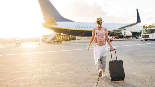 A traveler with a suitcase walks towards an airplane at sunset, wearing a straw hat and a backpack.