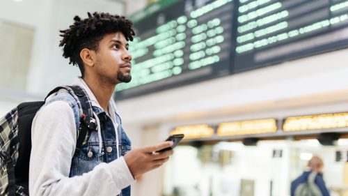 A young man with curly hair looks up at a departure board while holding a smartphone in an airport.