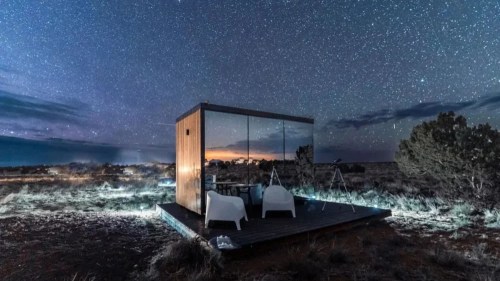 A modern cabin under a starry night sky, surrounded by desert landscape and illuminated by soft lights.