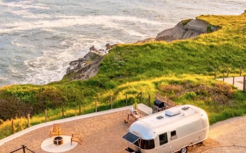A silver Airstream trailer parked on a grassy cliff overlooking the ocean, with chairs and a fire pit nearby.