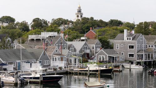 A scenic harbor with boats, quaint houses, and a clock tower in the background, surrounded by trees.