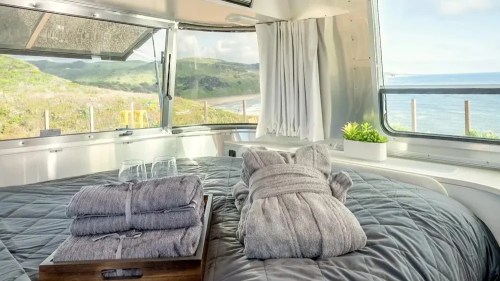 Cozy bedroom in a modern Airstream with a view of the ocean, featuring a neatly made bed and soft towels on a tray.