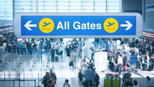 Sign directing travelers to "All Gates" at a busy airport, with a blurred crowd in the background.