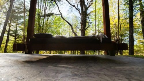 A wooden platform bed under a tree, surrounded by lush greenery and sunlight filtering through the leaves.