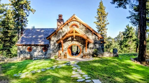 A charming stone cottage with a circular entrance, surrounded by trees and a stone pathway leading to the door.