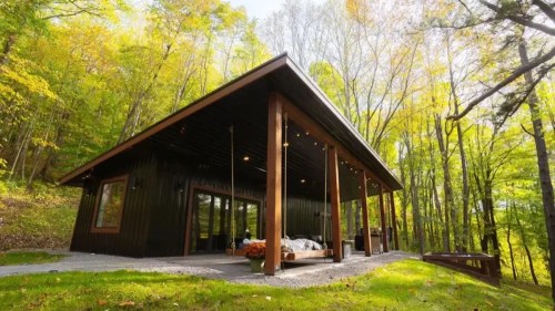 A modern black cabin surrounded by vibrant green trees and autumn foliage, featuring a spacious porch.