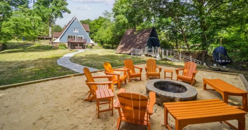 Large circular stone fire pit surrounded by orange chairs in a sandy area near the riverfront.