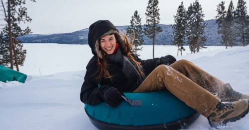 A woman in winter gear smiles as she rides on a blue tube down a snowy hill.
