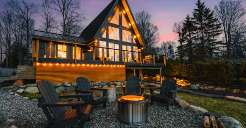 A large A-frame cabin with a firepit in front, illuminated by nighttime string lights.