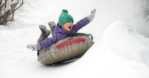 A child in winter gear rides on a snow tube, squealing with delight.