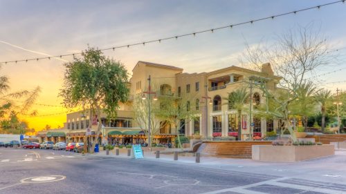A vibrant sunset illuminates a charming building with outdoor seating, surrounded by trees and a quiet street.