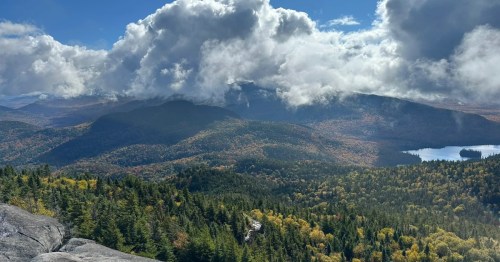 A wide vista of the view from the top of a mountain, with forests and clouds in the distance.