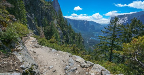 A narrow dirt trail winds along the side of a mountain, and Yosemite Valley sits far below.