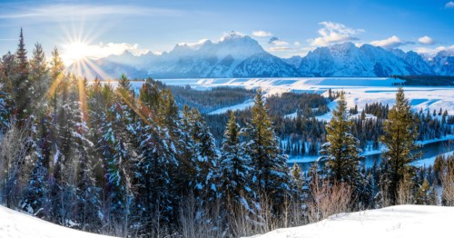 The sun peaks over the Snake River and a snowy forest in Grand Teton National Park.