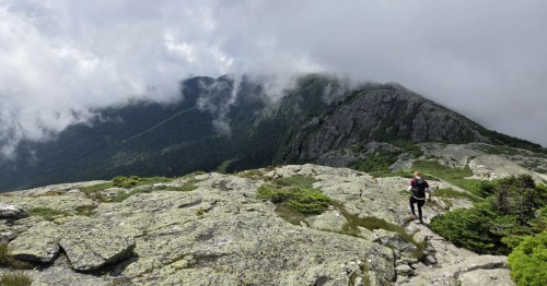 A hiker traverses an exposed rocky ridge on Mount Mansfield as clouds roll over the green mountain peaks.