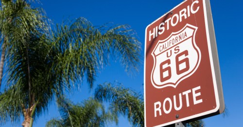 A brown Historic Route 66 road sign with palm trees in the background.