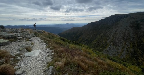 A hiker stands on a rocky overlook with a wide view of the rugged White Mountains valley and ridge.