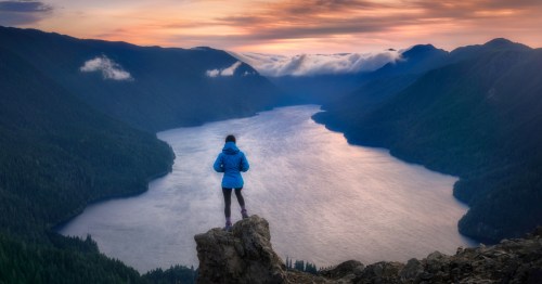 A woman stands on a tall rock, overlooking a large lake at sunset in Olympic National Park.