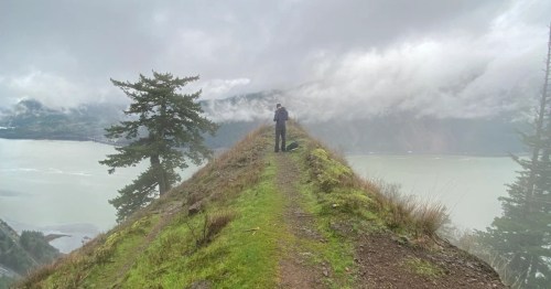 A hiker stands in the middle of a narrow trail with steep drop-offs on each side. Clouds hang low in the distance, and the Columbia River Gorge is visible.