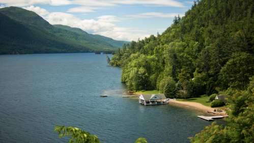 A serene lake surrounded by lush green mountains, featuring a small beach and a cozy cabin by the water's edge.