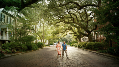 A couple walks hand in hand down a tree-lined, brick street, surrounded by lush greenery and historic buildings.