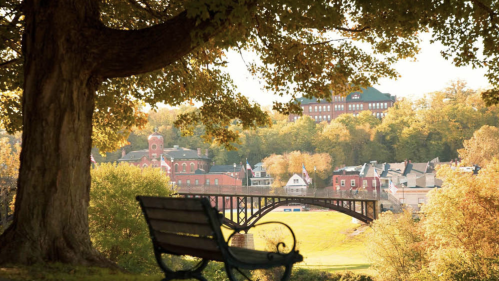 A scenic view of a town with a bridge, framed by trees and a bench in the foreground, showcasing autumn colors.