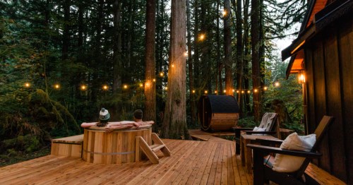 A couple sits in a cedar hot tub behind a cabin and next to a barrel sauna. String lights hang overhead, and tall trees surround them.