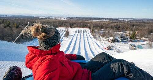 A woman in a red winter coat sits in a snow tube, looking down a hill covered in groomed snow tubing lanes.