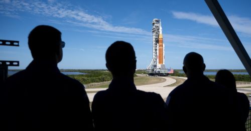 Silhouette of four people looking out at the massive Artemis II rocket being transported to the launch pad at Kennedy Space Center.