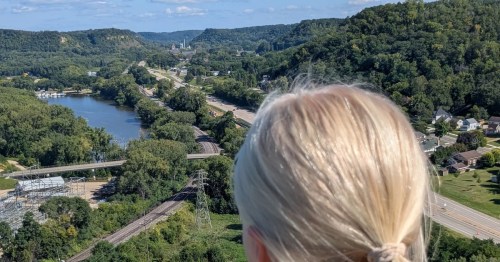 A blonde hiker looks out over the Mississippi River valley and the city of Red Wing from the scenic summit of Barn Bluff on a sunny day.