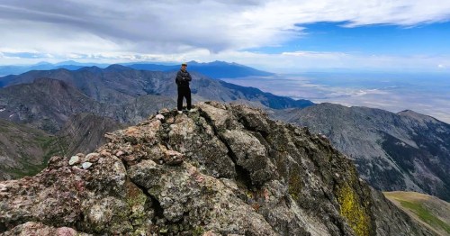 A hiker standing on the edges of the rocky terrain along Crestone Peak, an extreme fourteener hike in Colorado, with views of a vast valley and mountains in the distance.
