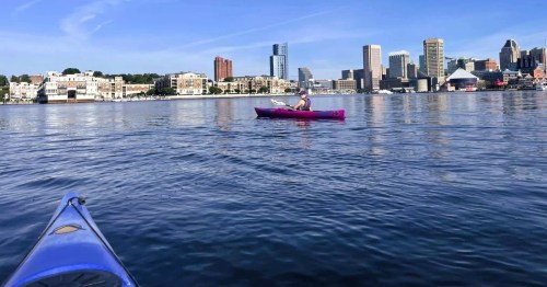 Two kayakers on a river along a paddle trail in Maryland with views of the Baltimore Harbor skyline in the distance.