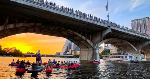 A group of kayakers on a sunset tour in Texas, drifting under a crowded bridge with the downtown Austin skyline in the distance.