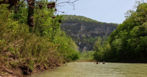 Kayakers paddling down a calm green river beneath towering limestone bluffs and lush spring foliage in the Arkansas Ozarks.