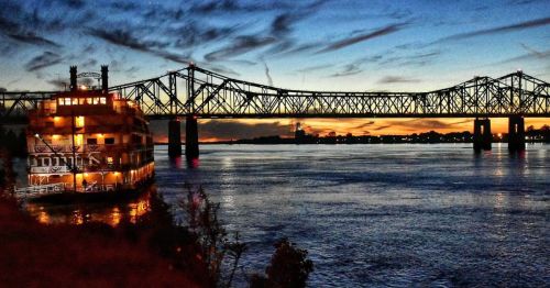 An illuminated steamboat docked near a bridge at sunset in Natchez, Mississippi.