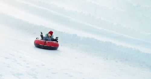 A happy child raises their hands in excitement while sliding down a groomed white slope in a red and black snow tube.