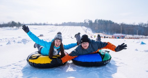An adult man and an adult woman ride snow tubes down a mountain on their bellies with huge grins on their faces.