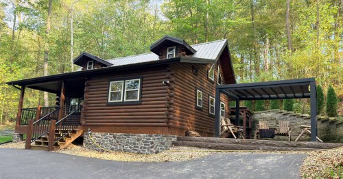 A dark wood log cabin with a metal roof tucked into a lush green forest with a stone foundation.