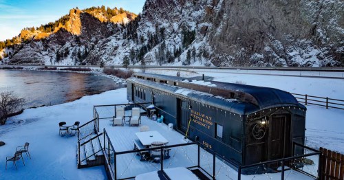 Exterior deck of a vintage railcar Airbnb in winter with snow-covered seating, string lights, and a river flowing beside rocky canyon walls.