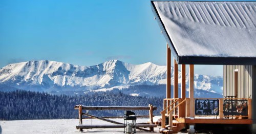 Snow-covered cabin porch with wooden railing facing dramatic, snow-capped mountains under a bright blue winter sky.