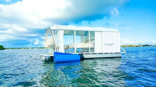 A floating suite on calm water, featuring a white canopy and blue accents, under a bright blue sky with clouds.