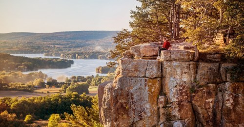 A hiker sits on the edge of a sheer limestone cliff at Gibraltar Rock State Natural Area in Wisconsin, overlooking the expansive Wisconsin River valley and Lake Wisconsin during golden hour.